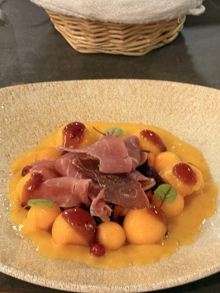 A starter of melon balls and thin slices of cured ham in an orange sauce